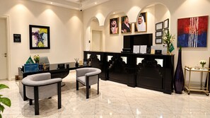 A reception area with a black desk, two grey chairs, a marble floor, framed portraits on the wall, and a large abstract painting.