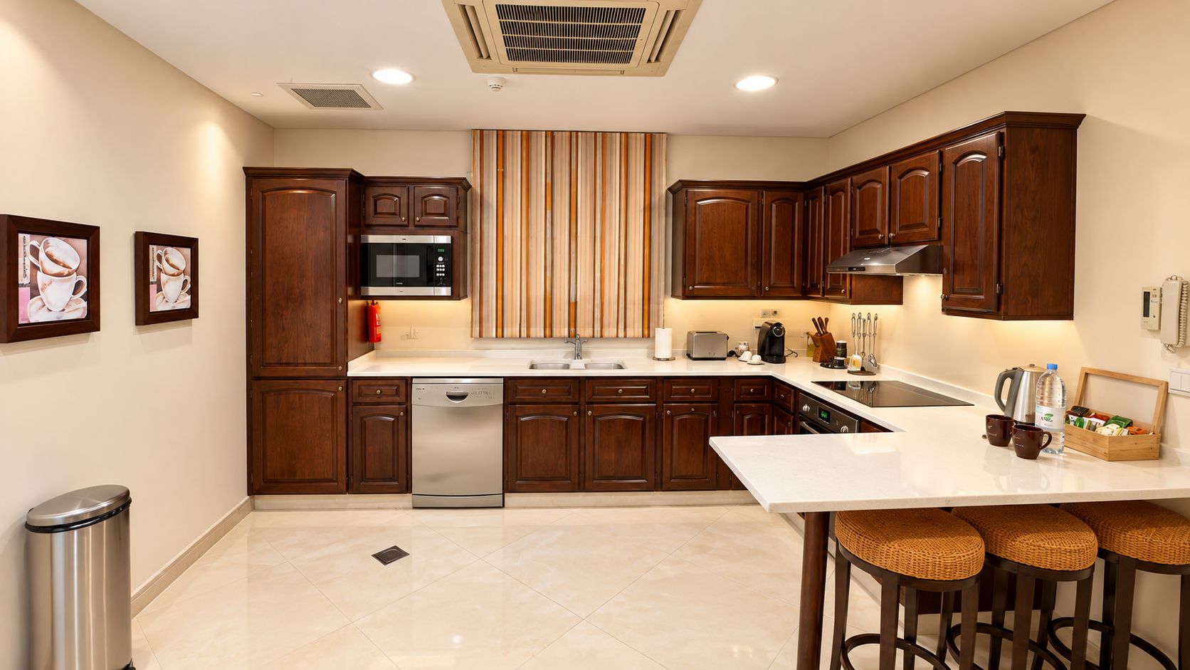 A modern kitchen with wooden cabinets, a stainless steel dishwasher, a microwave, and a sink. There is a bar area with stools and a small table with a coffee machine and cups.