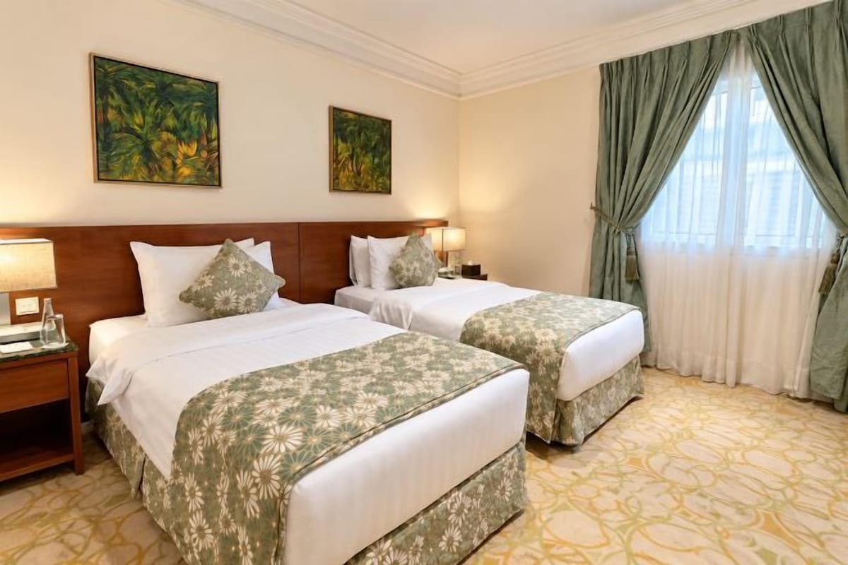 A hotel room with two beds, a wooden headboard, a nightstand with a lamp, and a window with curtains.