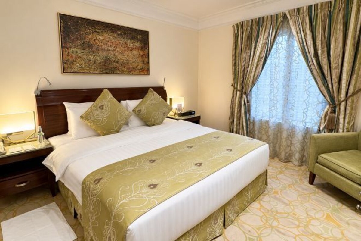 A hotel room with a large bed, a nightstand, a green armchair, and a window with curtains.