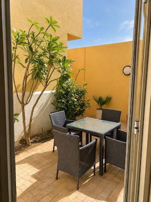 Un patio exterior con una mesa de vidrio y sillas de mimbre, rodeado de plantas en macetas y una pared amarilla.