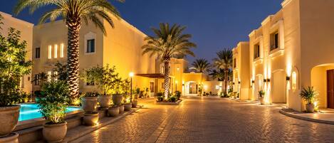A courtyard with a swimming pool, potted plants, and buildings with warm lighting.