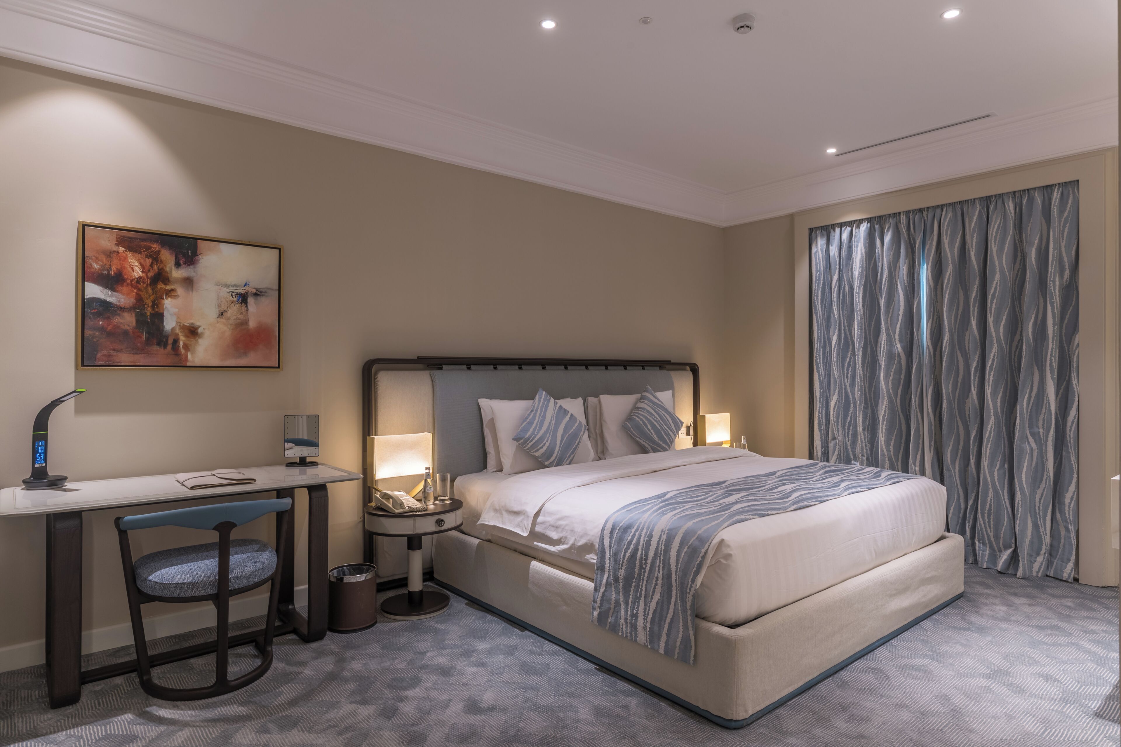Habitación de hotel con cama, escritorio, silla, lámparas de noche y un cuadro en la pared.