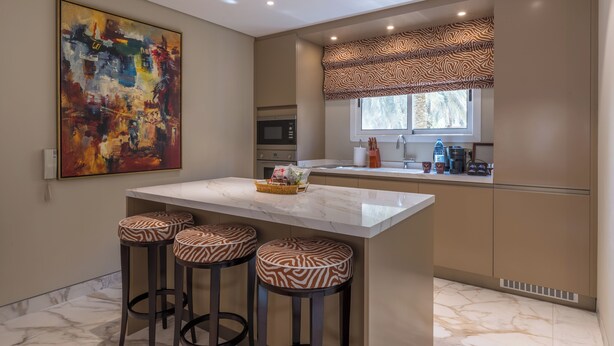 Een moderne keuken met een marmeren kookeiland, barstoelen, een groot abstract schilderij en een raam met een patroon gordijn.