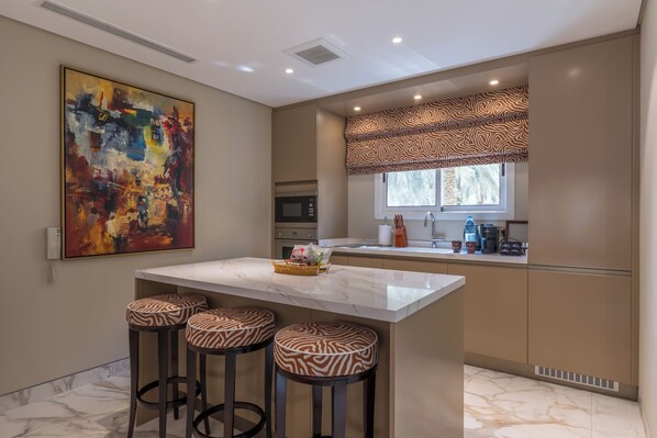 A modern kitchen with a marble island, bar stools, a large abstract painting, and a window with patterned valance.