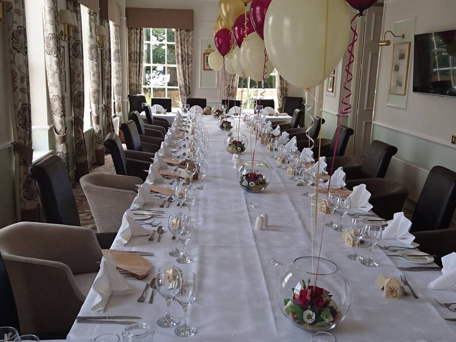 A long dining table set for a formal event, with chairs on both sides and balloons in the background.