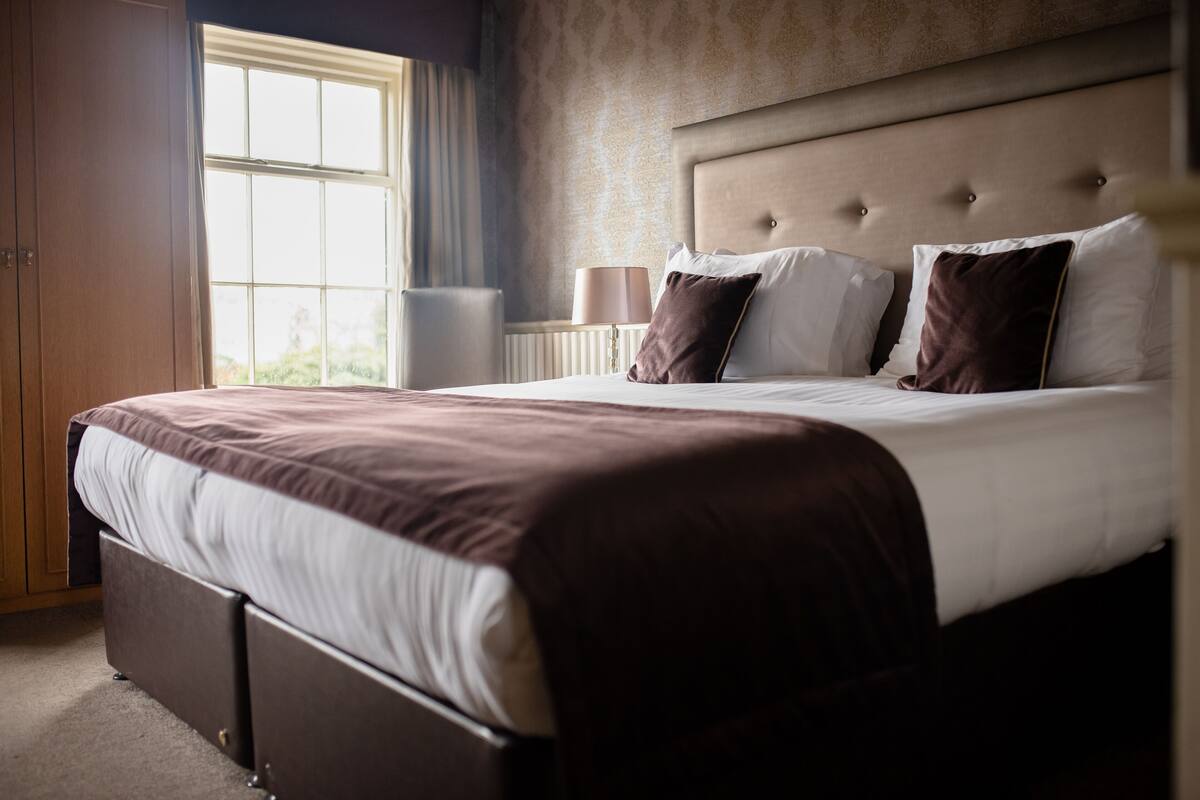 A neatly made bed with a headboard, a bedside lamp, and a window with curtains.