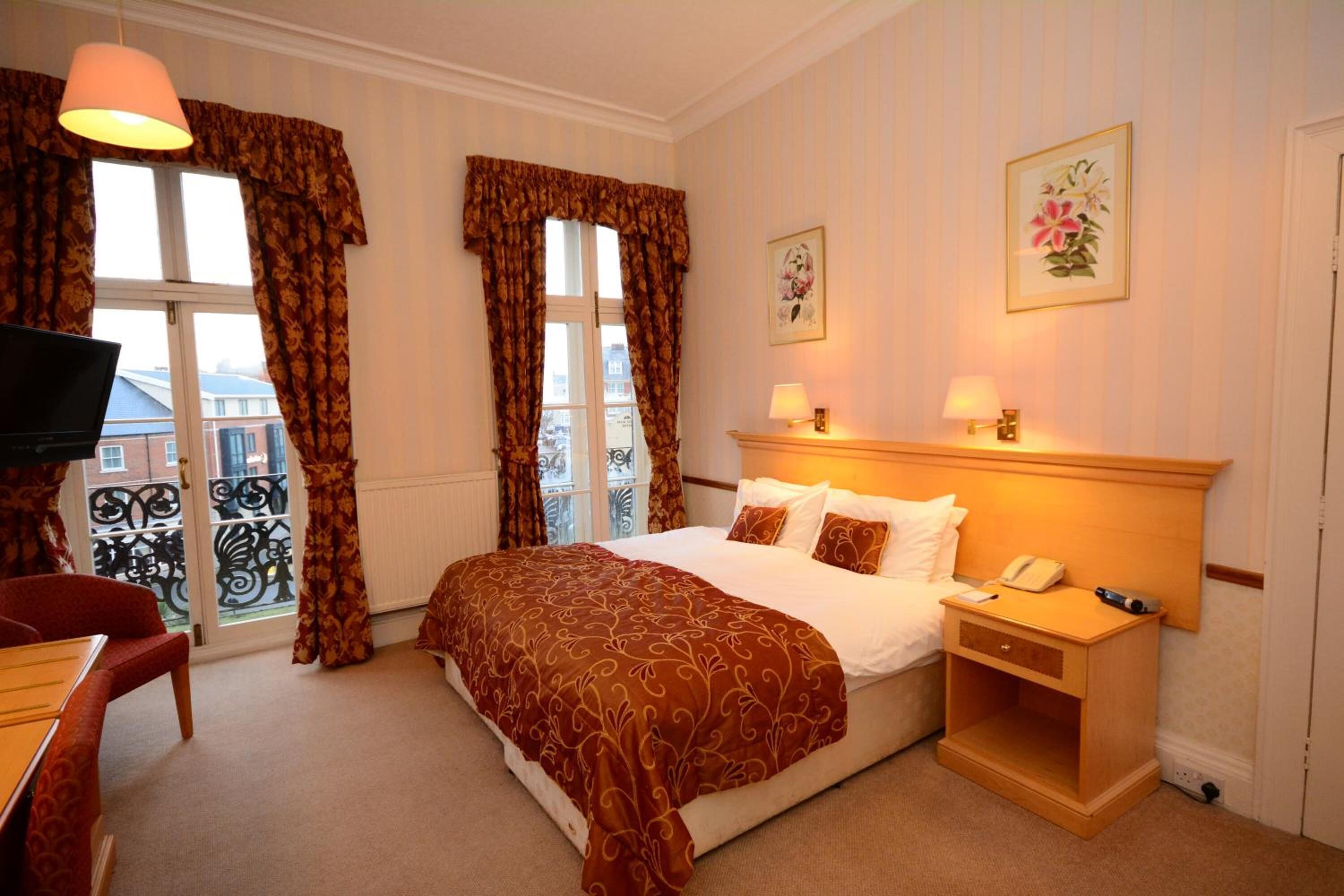 A hotel room with a bed, bedside table, lamp, and a TV on a desk.