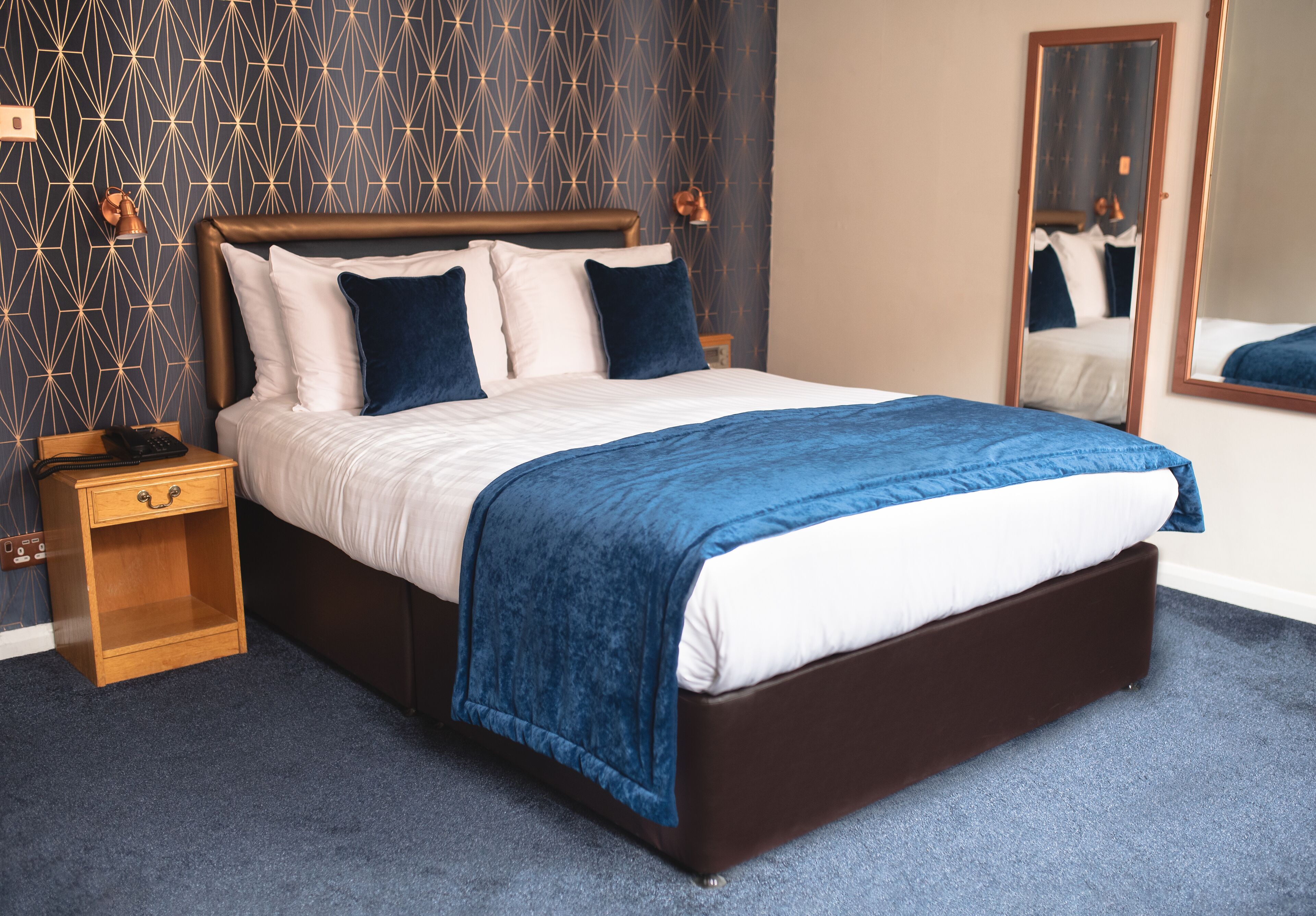 A neatly made bed with a blue blanket, a wooden nightstand with a phone, and a patterned wallpaper.