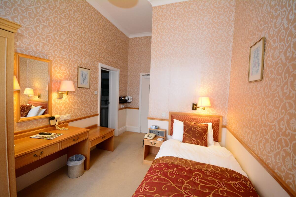 A hotel room with a bed, bedside table, lamp, mirror, and a desk with a telephone and trash bin.