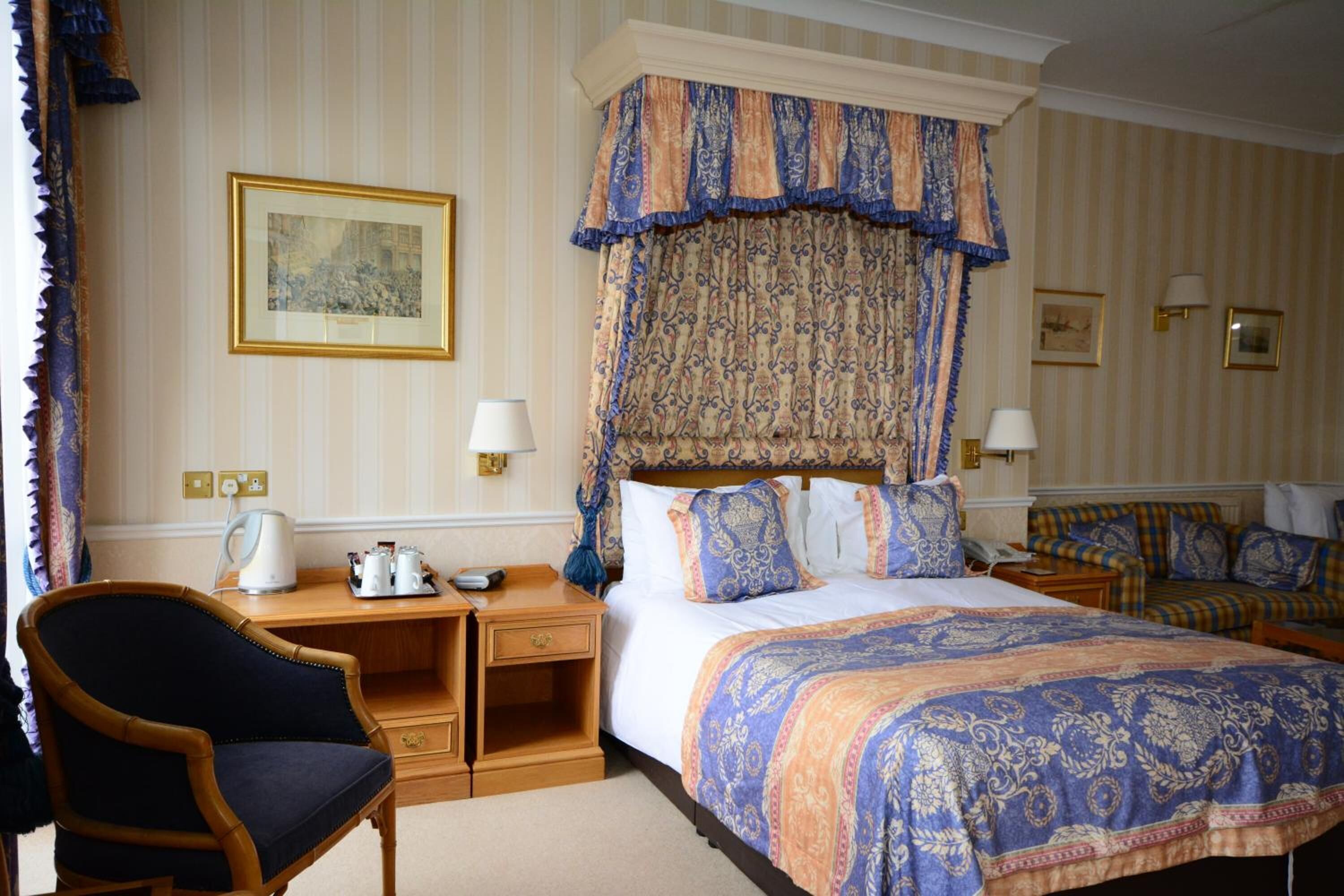 A hotel room with a large bed, a desk with a chair, a bedside table with a lamp, and a wall-mounted telephone.