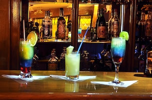 Un bar con tres bebidas sobre la barra, varias botellas de licor al fondo, y un espejo que refleja la zona del bar.