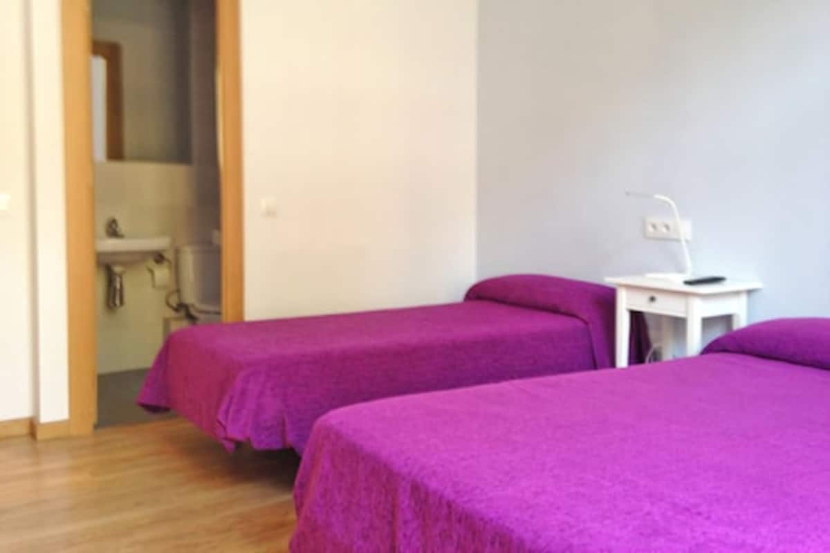 A room with two beds covered in purple bedding, a white bedside table with a lamp, and a bathroom visible through an open door.