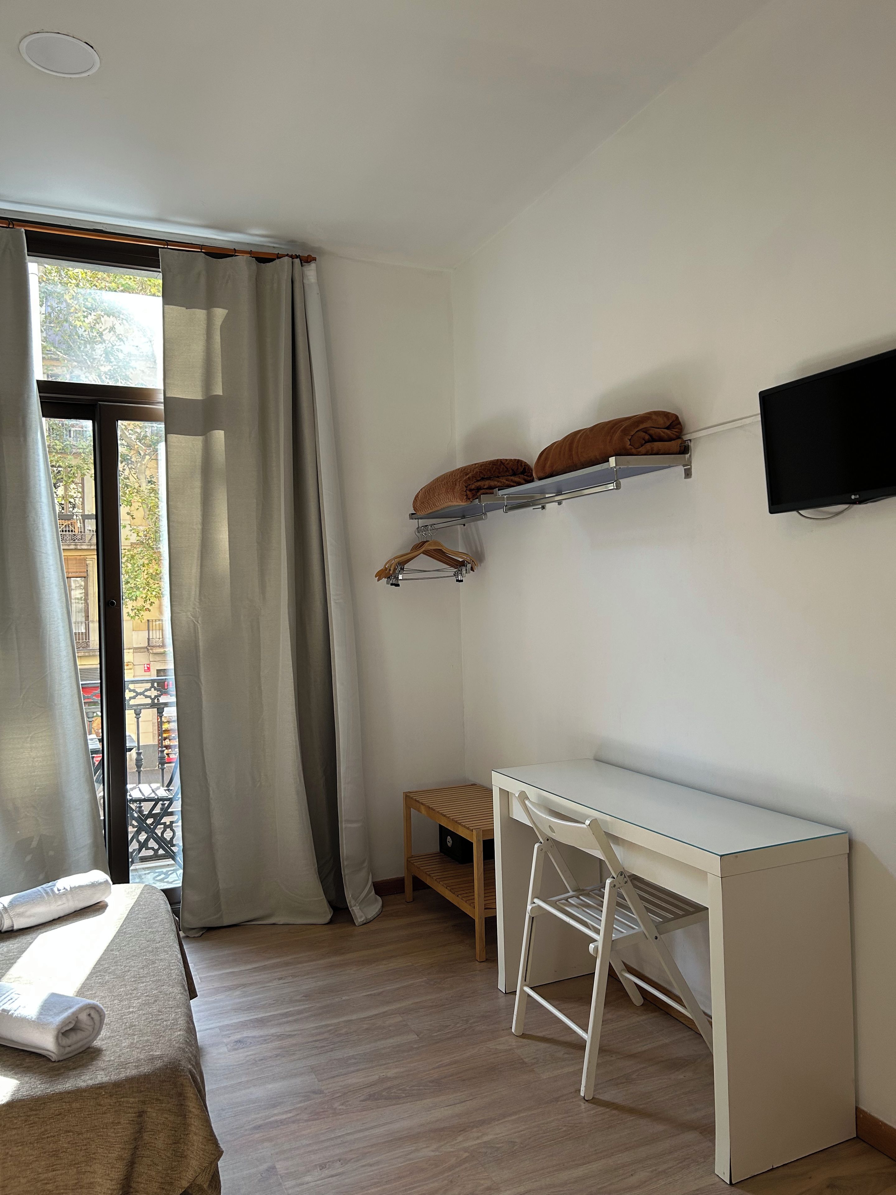 Una habitación con cama, escritorio, televisor montado en la pared y ventana con cortinas.