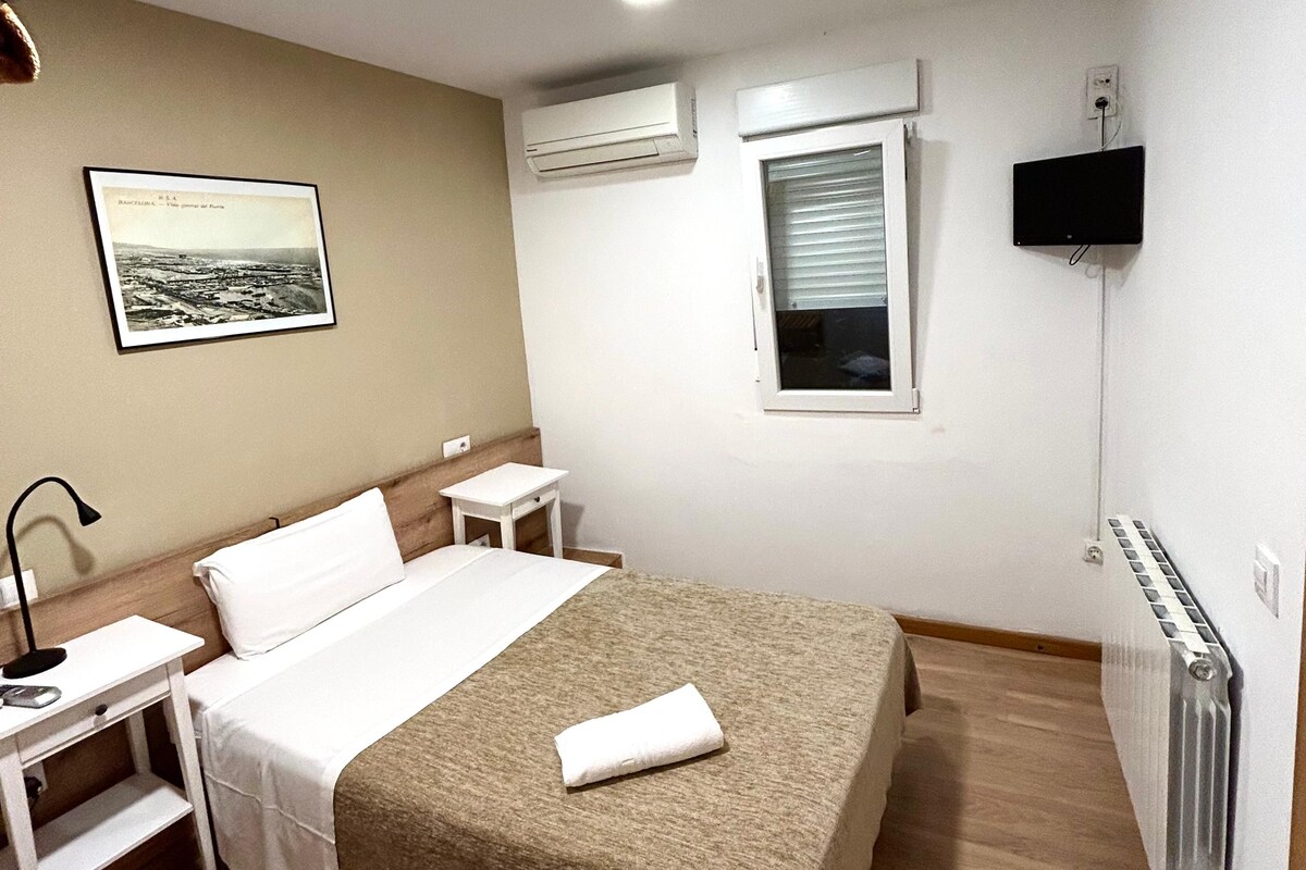 A hotel room with a bed, bedside tables, a lamp, a picture on the wall, an air conditioning unit, a window, and a TV.