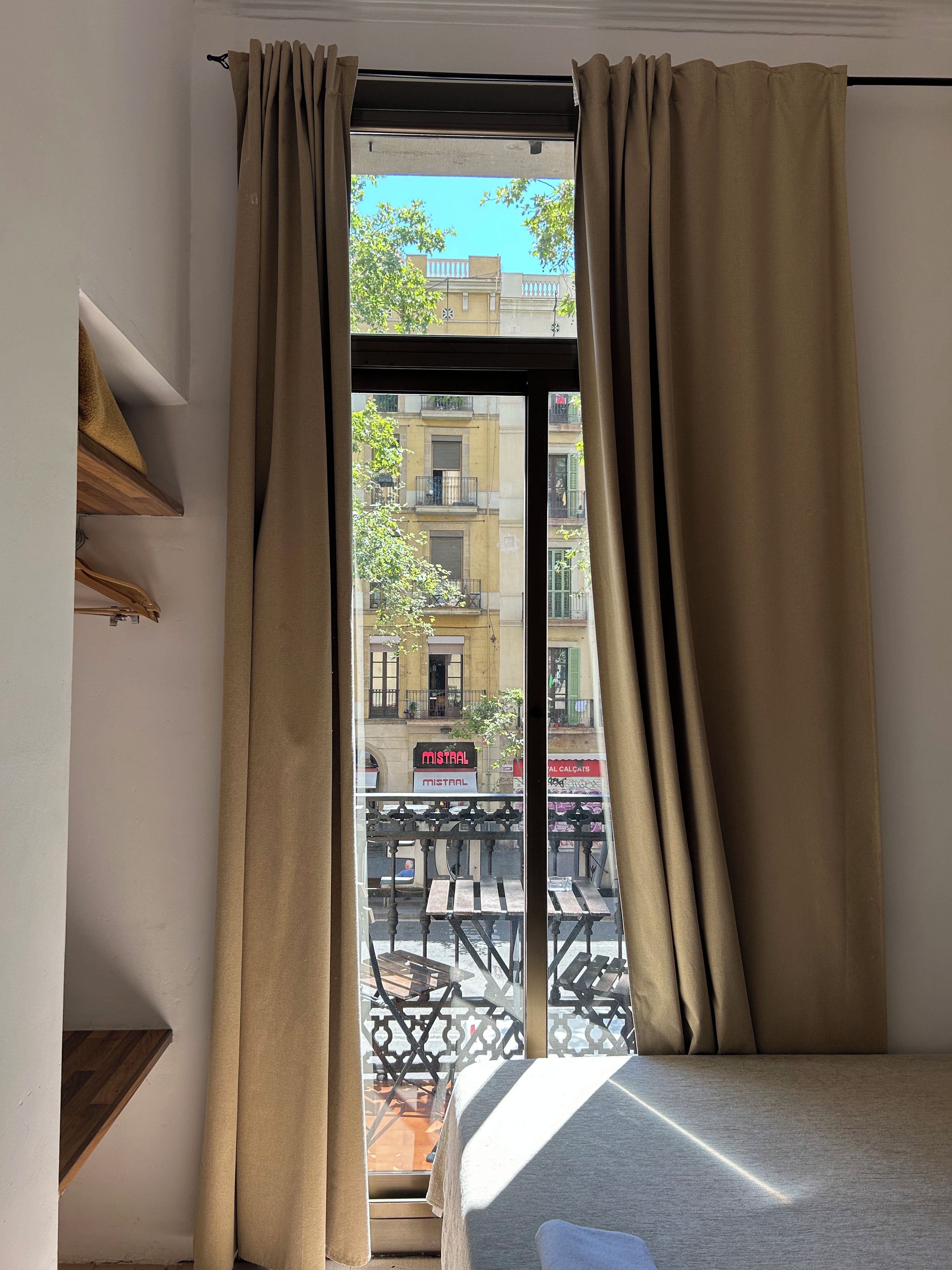 Una habitación con cama, ventana con cortinas y vista a la calle, con un letrero que dice 'MISTRAL'.