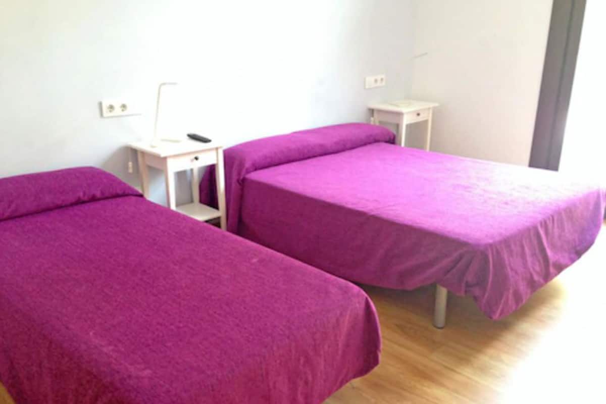 A room with two single beds, each with a purple bedspread. There is a small white nightstand with a lamp between the beds.