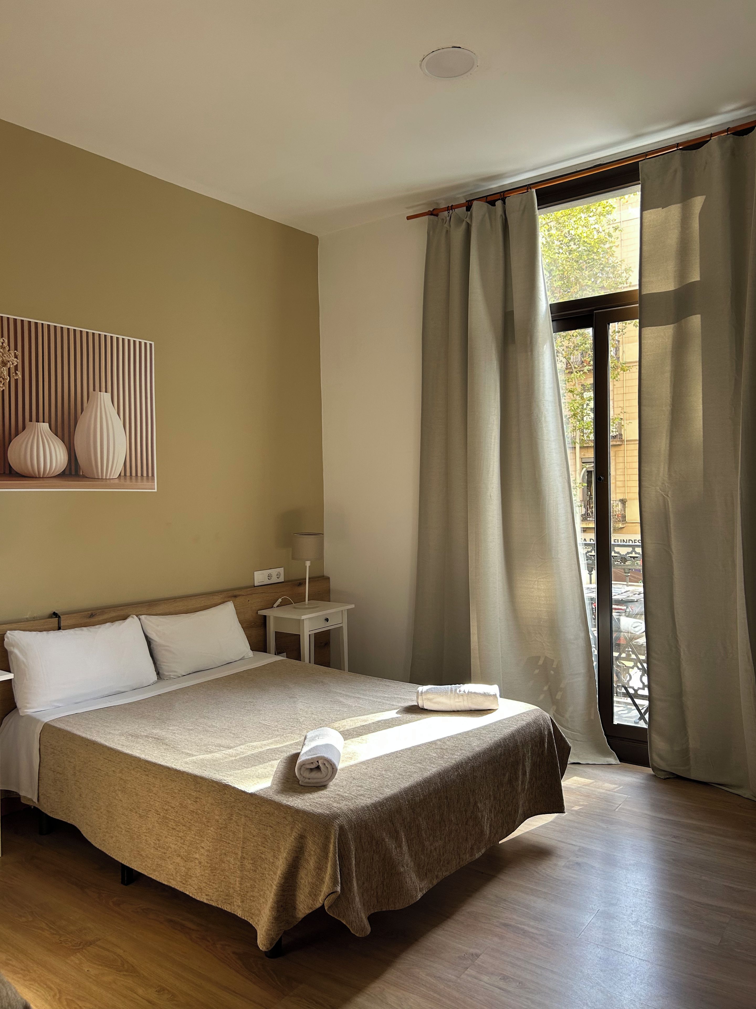 Habitación de hotel con cama, mesita de noche, lámpara y vistas a través de una ventana con cortinas.
