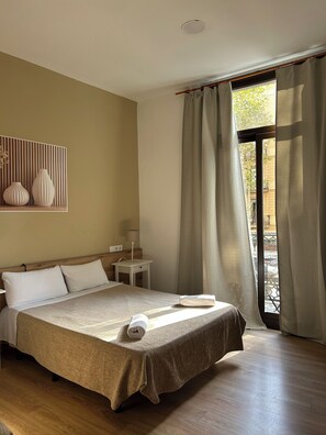 Habitación de hotel con cama, mesita de noche, lámpara y vistas a través de una ventana con cortinas.