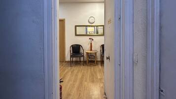 A hallway leading to a room with a wooden floor, a clock on the wall, and two chairs.