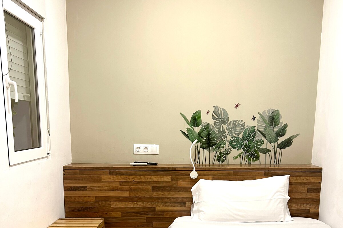 A hotel room with a wooden headboard, a bed with white bedding, a small wooden bench, a wall-mounted power outlet, and a wall decoration of tropical leaves.