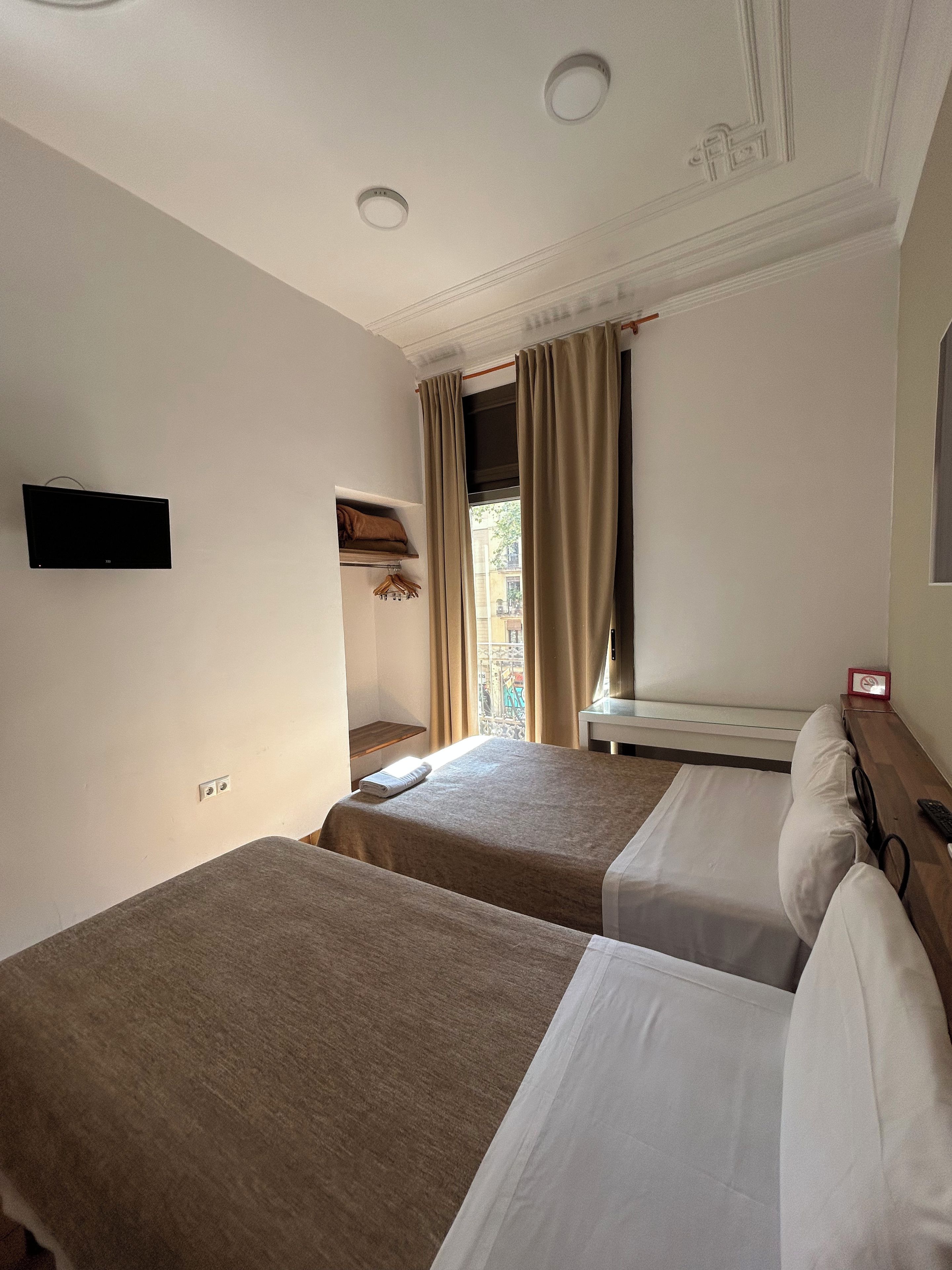 Habitación de hotel con dos camas, un televisor de pantalla plana montado en la pared, una mesita con un letrero rojo y una ventana con cortinas.