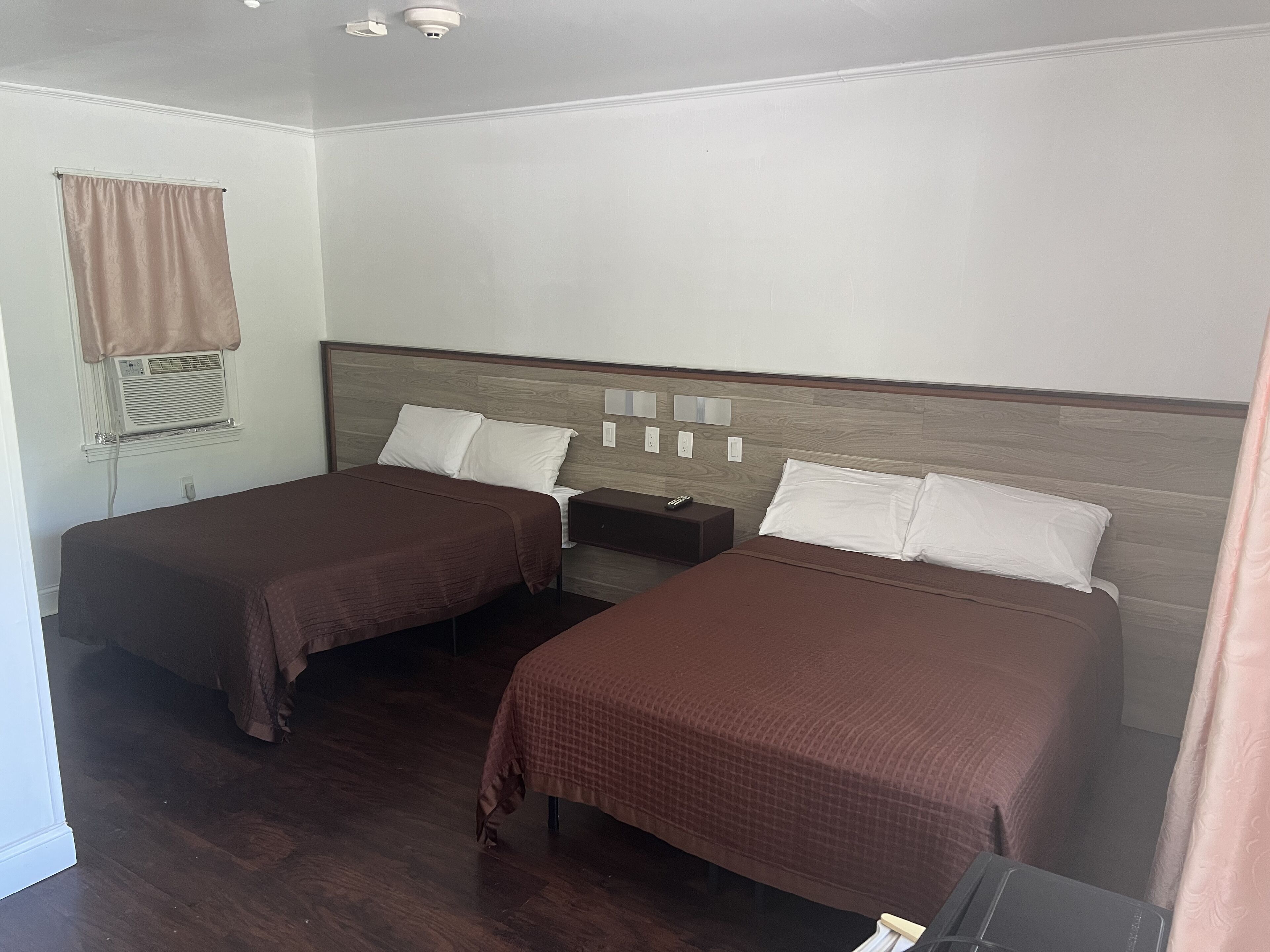 A hotel room with two beds, a nightstand, a window with a curtain, and a ceiling-mounted air conditioning unit.