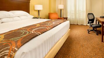 A hotel room with a large bed, two bedside lamps, an orange chair, a desk with a chair, and a window with curtains.
