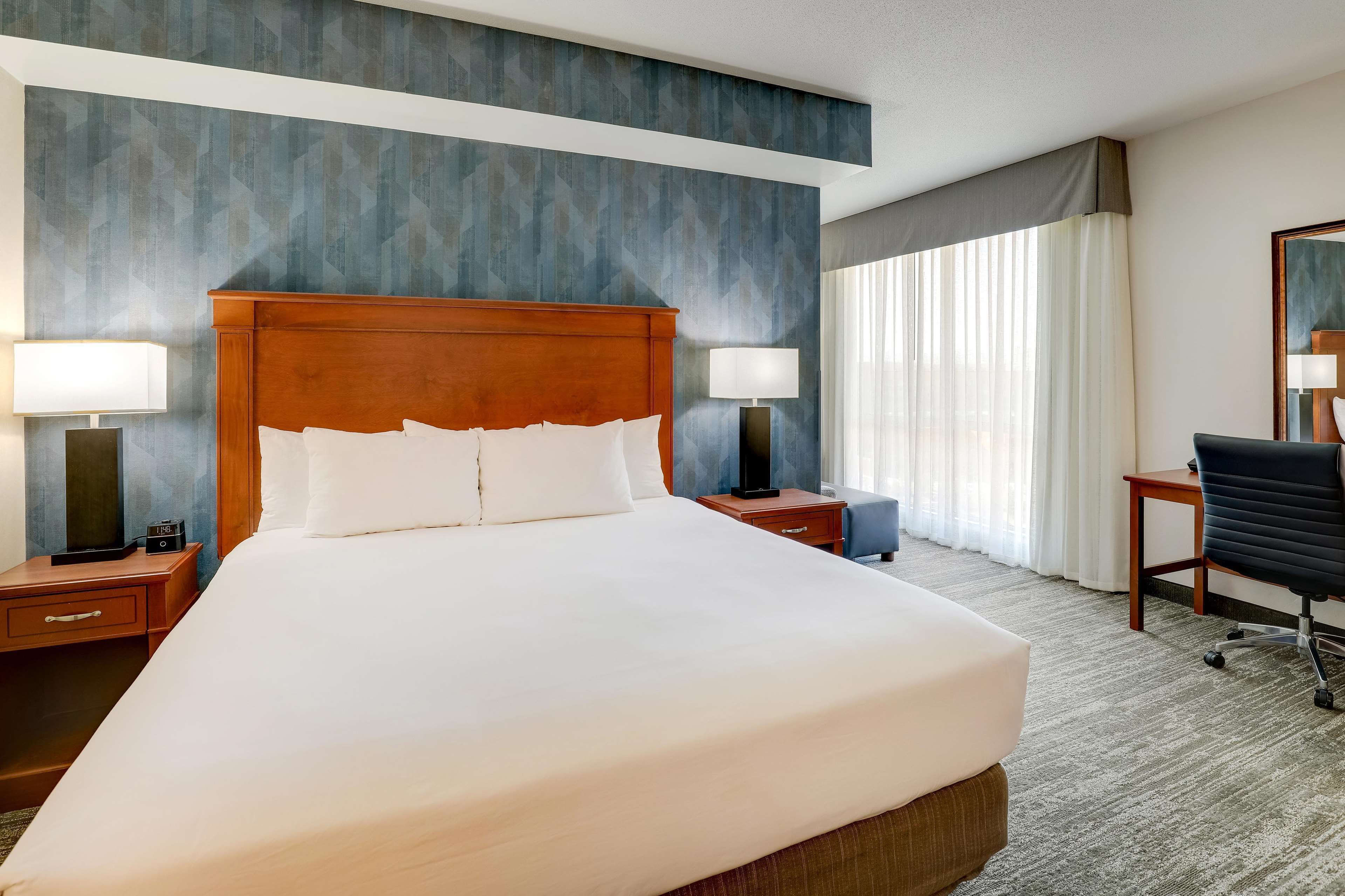 A hotel room with a large bed, two bedside tables with lamps, a desk with a chair, and a large window with curtains.