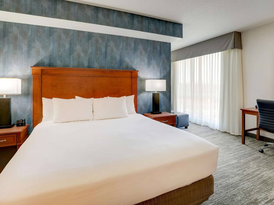 A hotel room with a large bed, two bedside tables with lamps, a desk with a chair, and a large window with curtains.