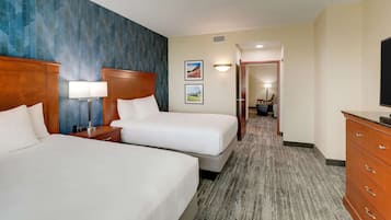 Quarto de hotel com duas camas, cabeceira de madeira, mesa de cabeceira com abajur, cômoda e corredor que leva a um banheiro.