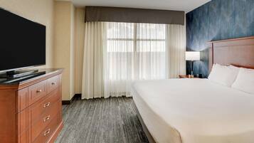 A hotel room with a large bed, a flat-screen TV, a wooden dresser, and a window with sheer curtains.