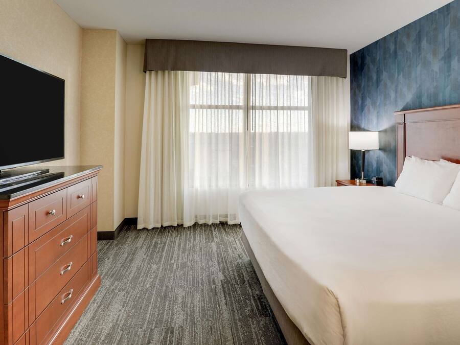 A hotel room with a large bed, a flat-screen TV, a wooden dresser, and a window with sheer curtains.