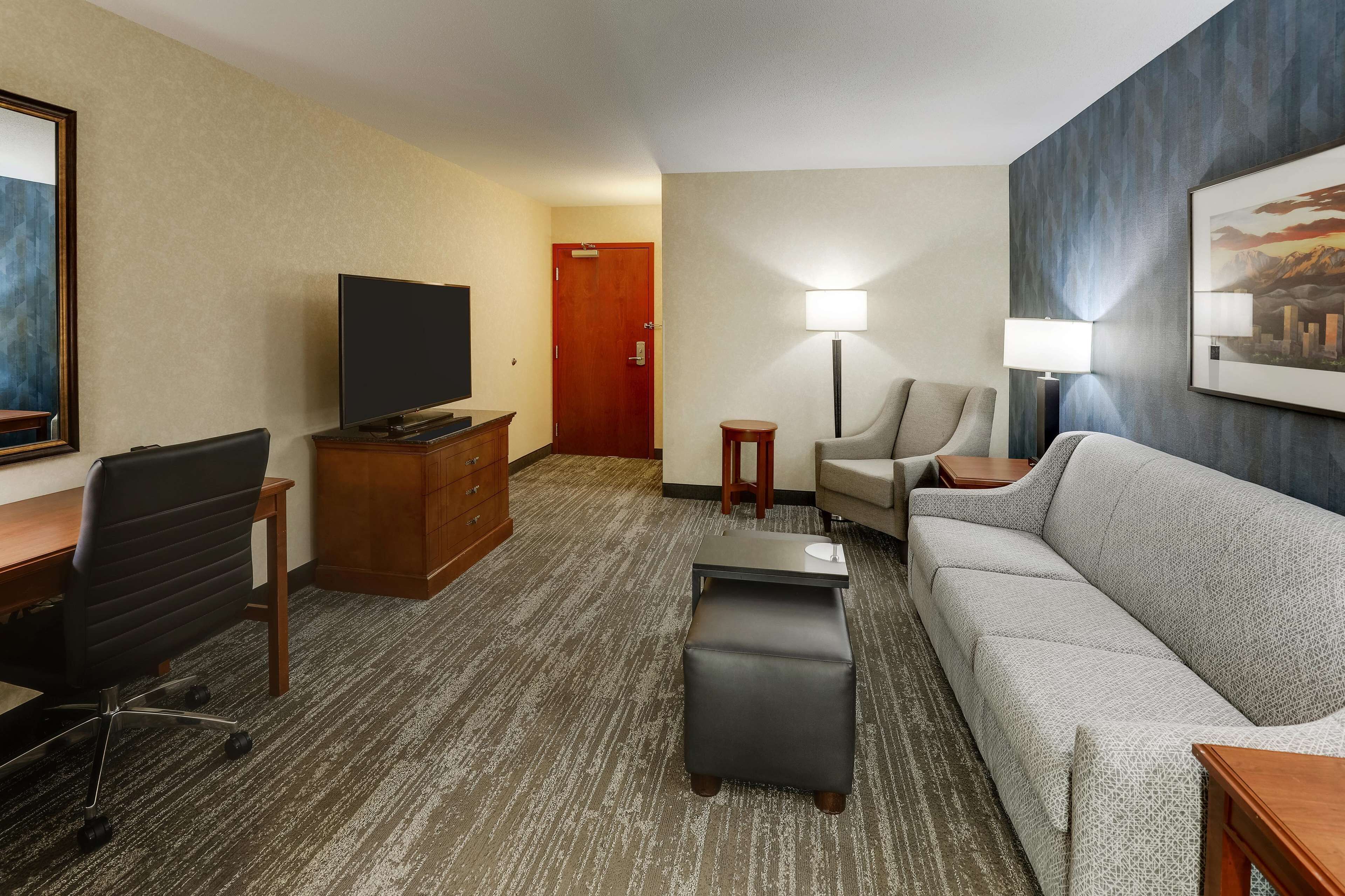 A hotel room with a flat-screen TV, a sofa, a desk with a chair, a small table, and a lamp.