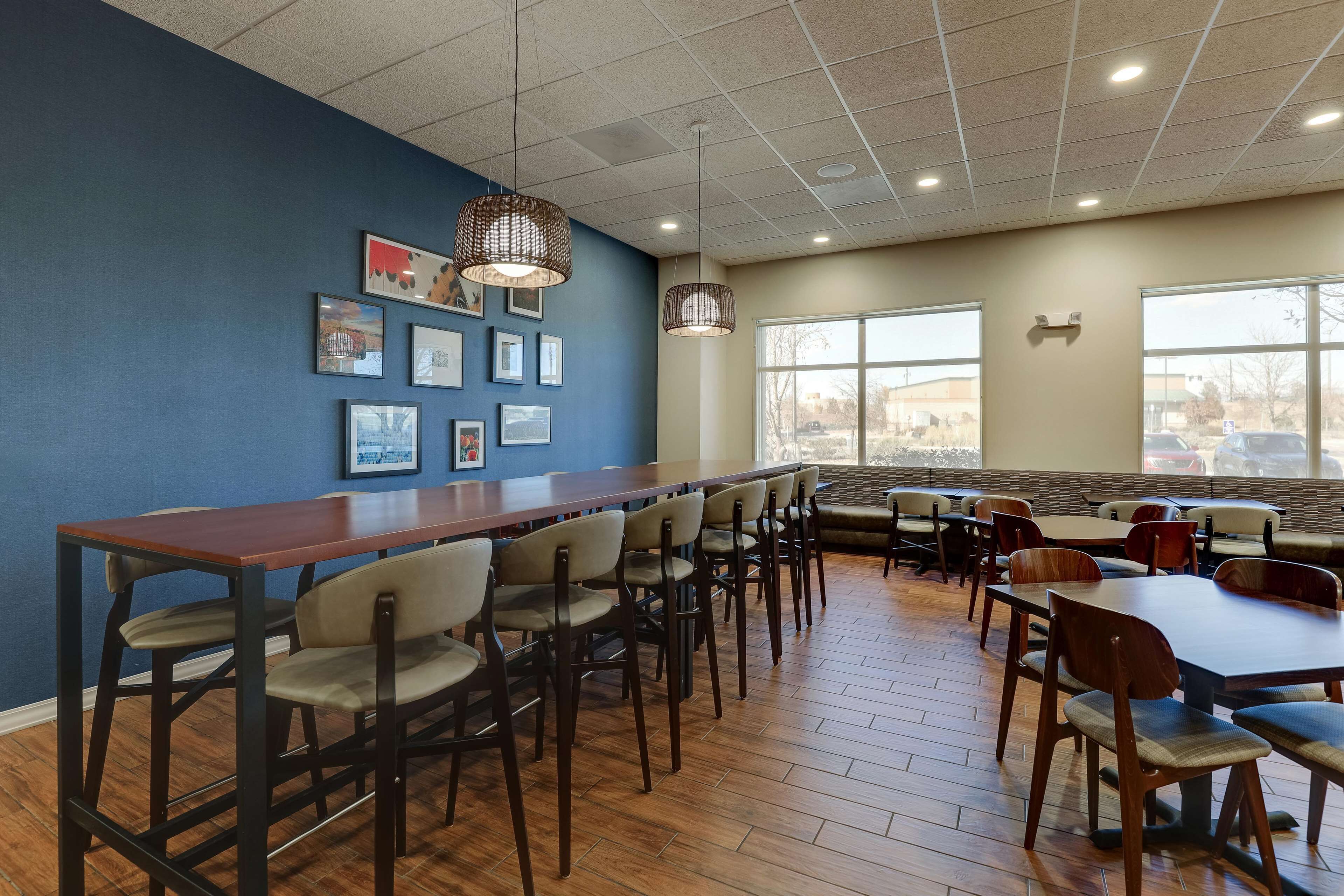 Un vaste espace dédié à la salle à manger, avec une longue table en bois et de nombreuses chaises, de grandes fenêtres offrant une vue sur l’extérieur, et des tableaux encadrés accrochés au mur bleu.