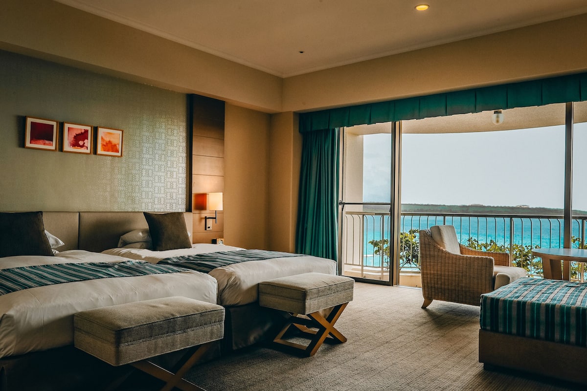 Habitación de hotel con dos camas, un balcón y vistas al mar, y una silla de mimbre.