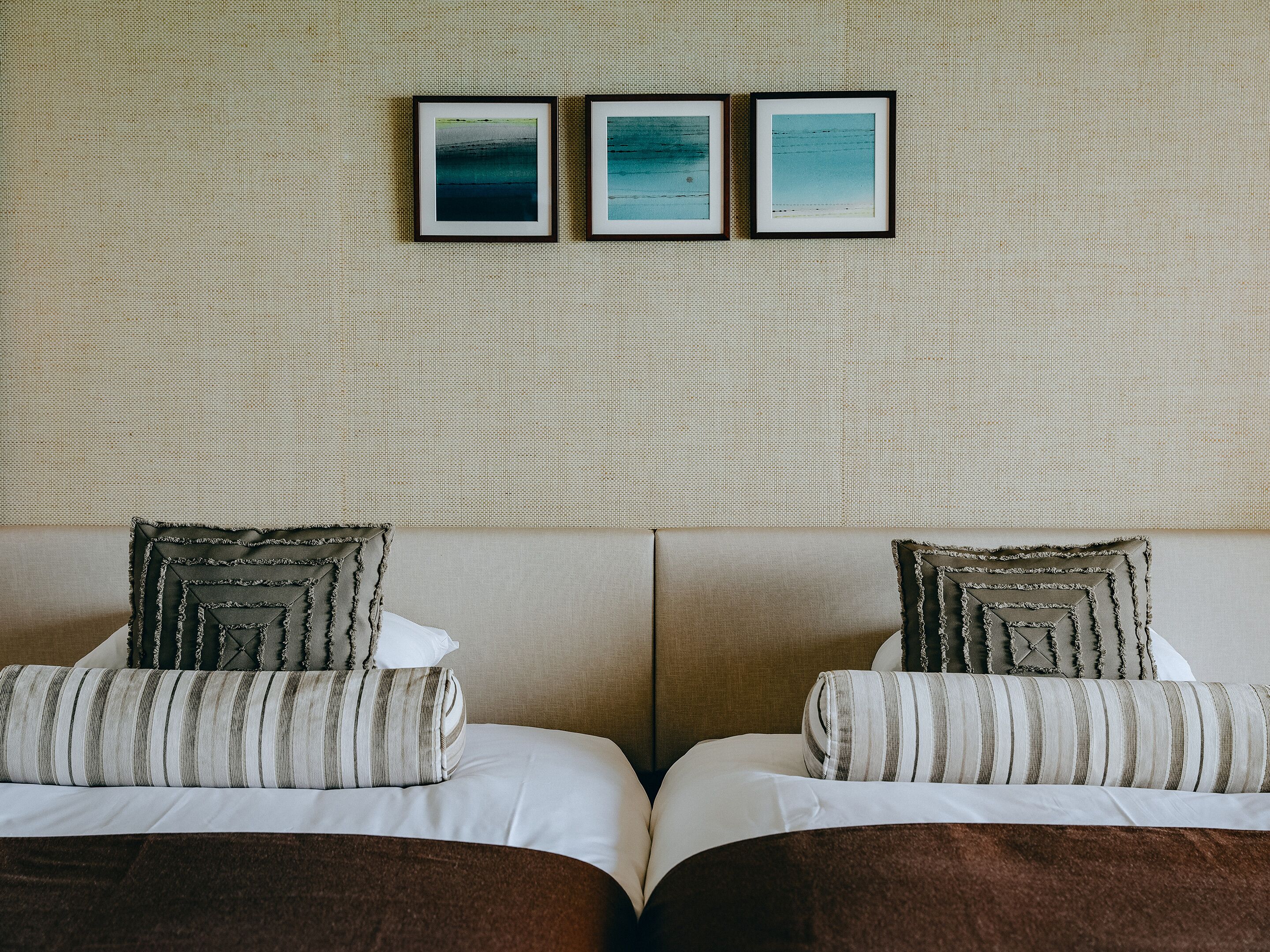 Habitación de hotel con dos camas, cada una con ropa de cama a rayas y almohadillas decorativas. Hay tres cuadros enmarcados en la pared sobre las camas.