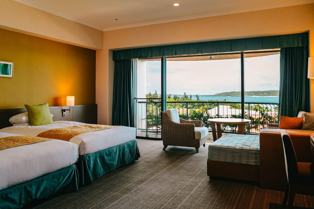 Habitación de hotel con dos camas, un balcón con vista al mar y una zona de estar con una mesa.
