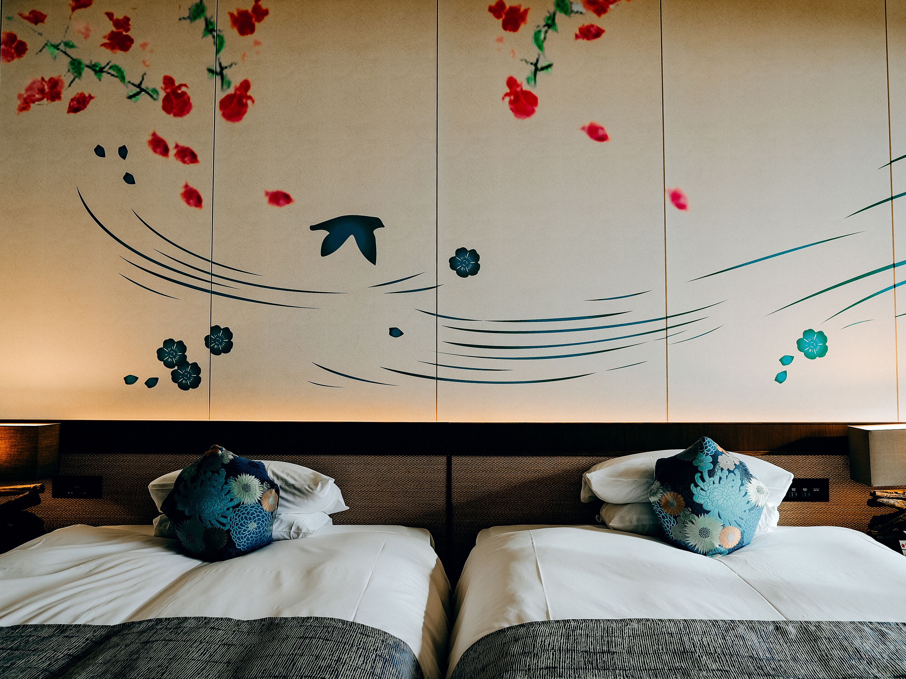 Ein Hotelzimmer mit zwei Betten, jedes mit einer weißen Matratze und einem gemusterten Kissen. Das Kopfteil ist dunkel und über den Betten befindet sich eine Wanddekoration.