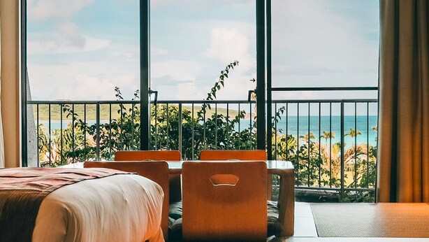 Habitación de hotel con un ventanal grande, una cama, una mesa con sillas y vistas a la playa y al océano.