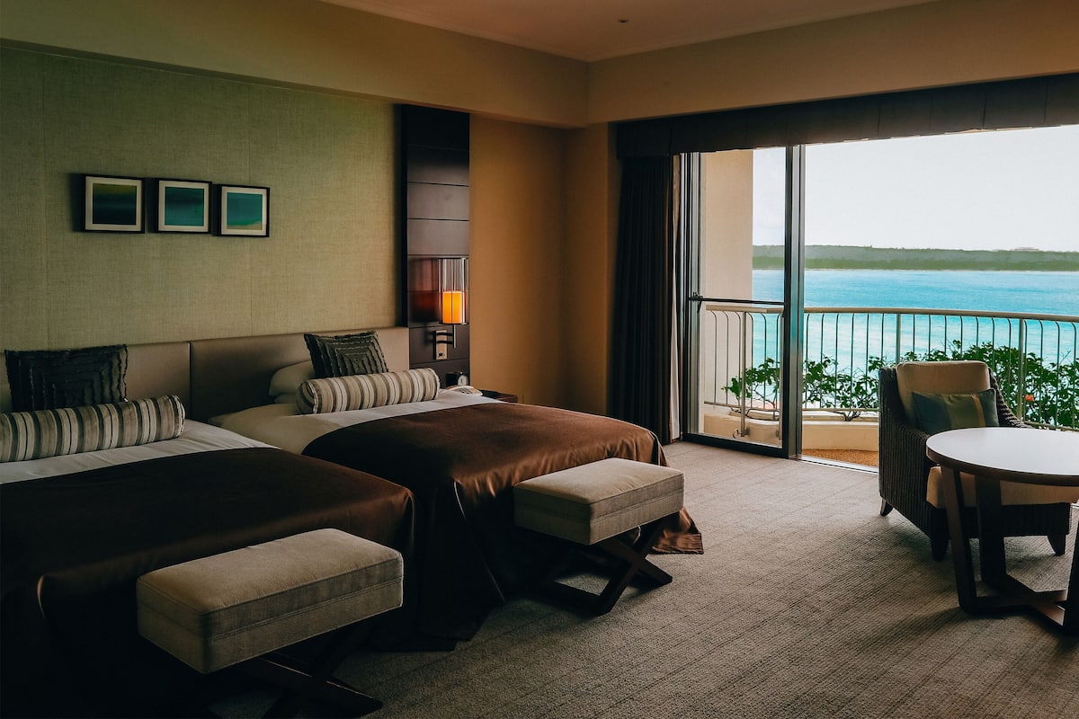 Habitación de hotel con dos camas, una mesita y una silla. Tiene un balcón con vista al mar.