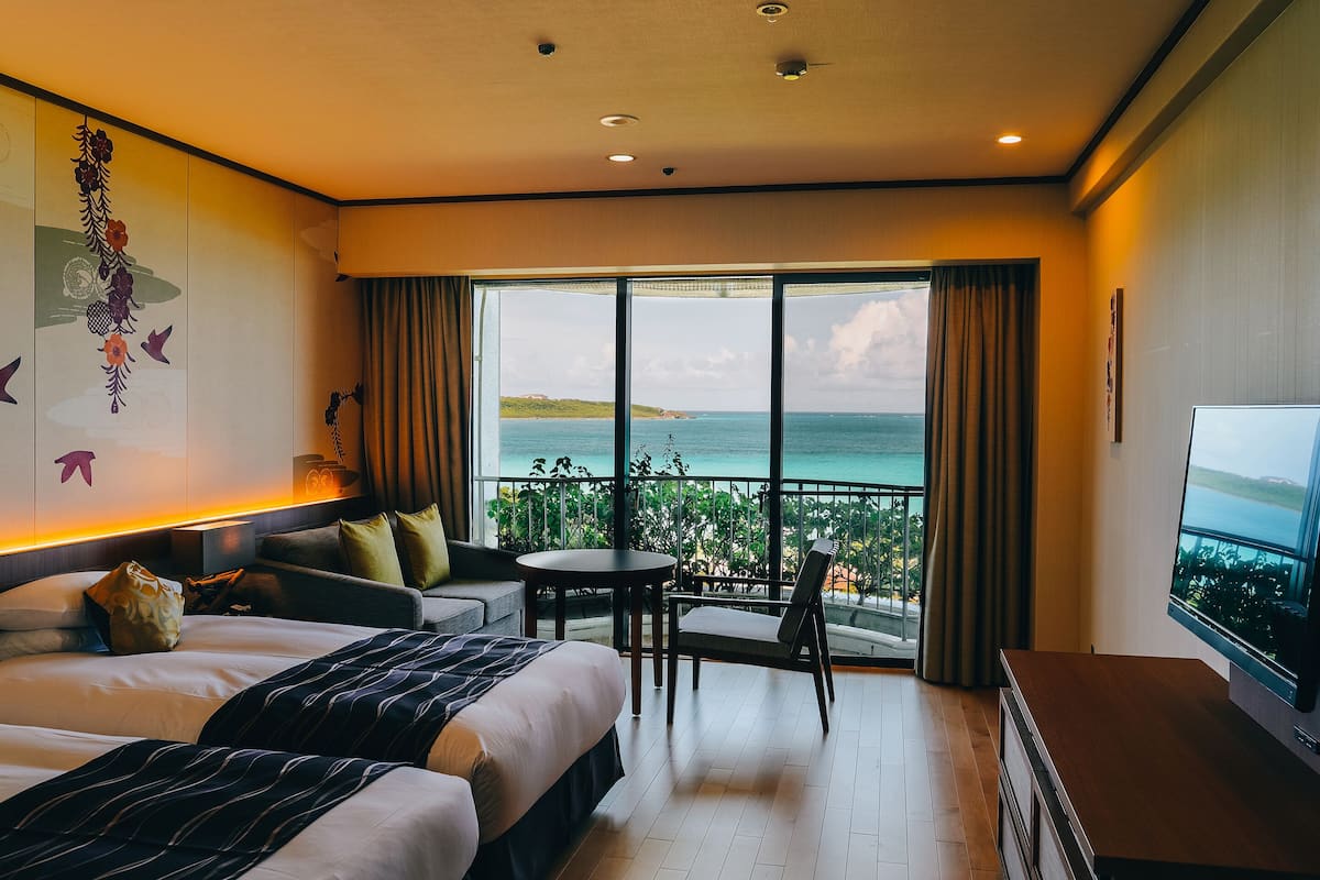 Habitación de hotel con una cama grande, un sofá, una mesita y un televisor. La habitación tiene un balcón con vistas al mar.