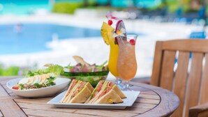 Una mesa con un plato de sándwiches, un vaso de bebida tropical y un bol de ensalada, con vista a una piscina y una playa.