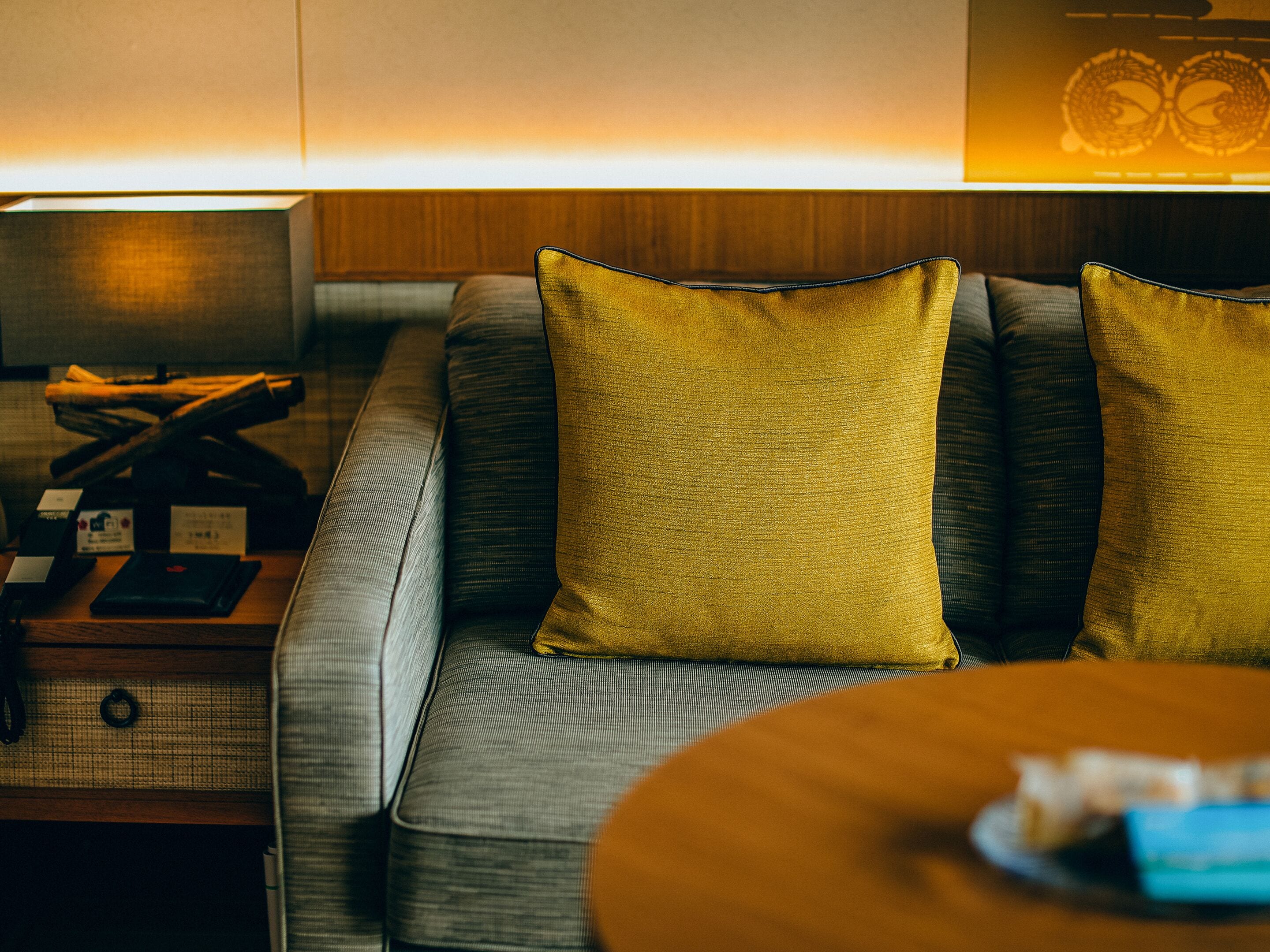 A modern hotel room with a sofa, two yellow pillows, a wooden side table, and a lamp.