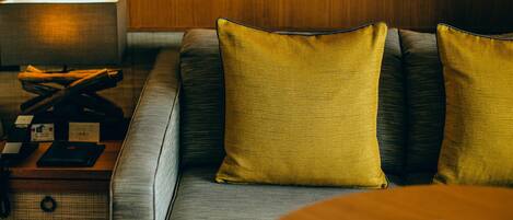 A modern hotel room with a sofa, two yellow pillows, a wooden side table, and a lamp.