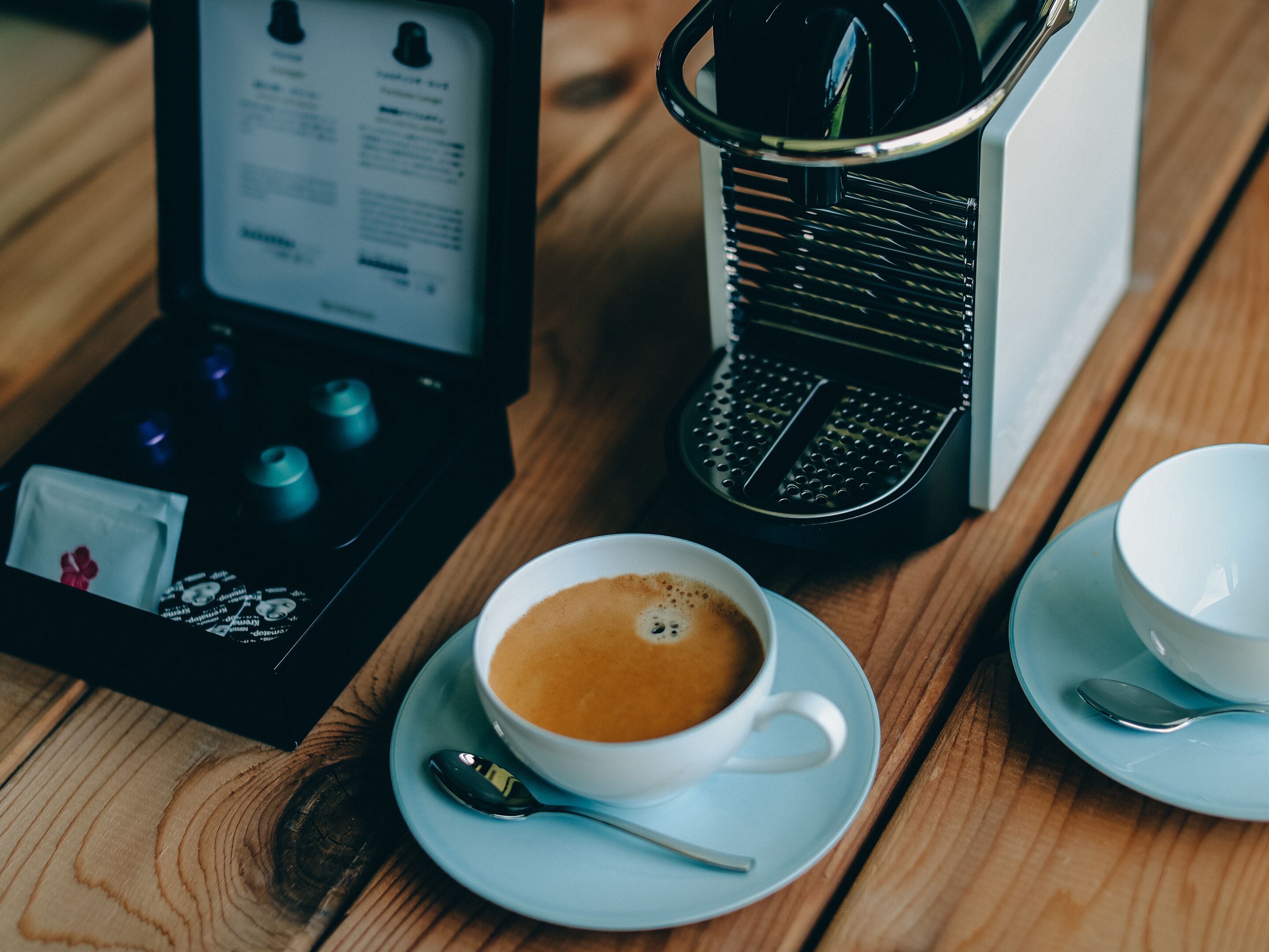 En kopp kaffe på ett litet fat med en tesked, en kaffebryggare och en ask med kaffekapslar på ett träbord.