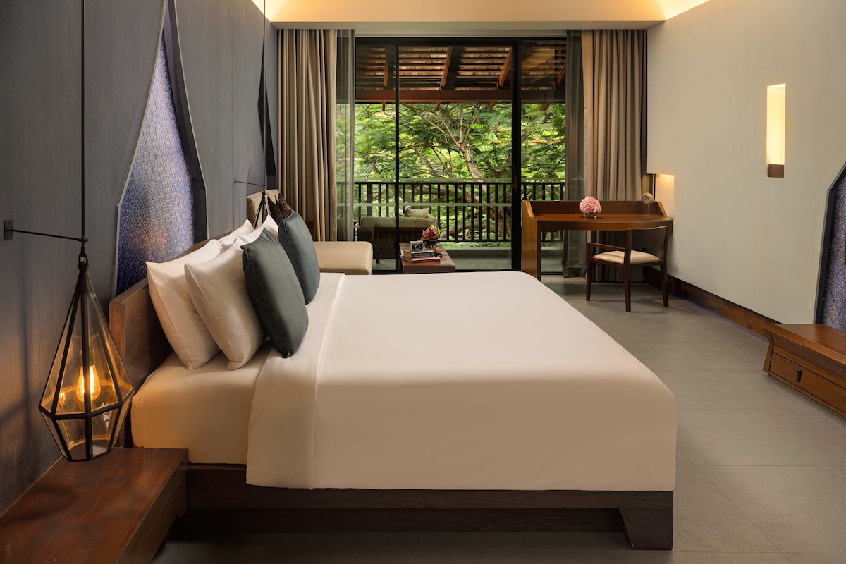 Habitación de hotel con una cama grande, un escritorio, una silla y un balcón con vistas a la vegetación.
