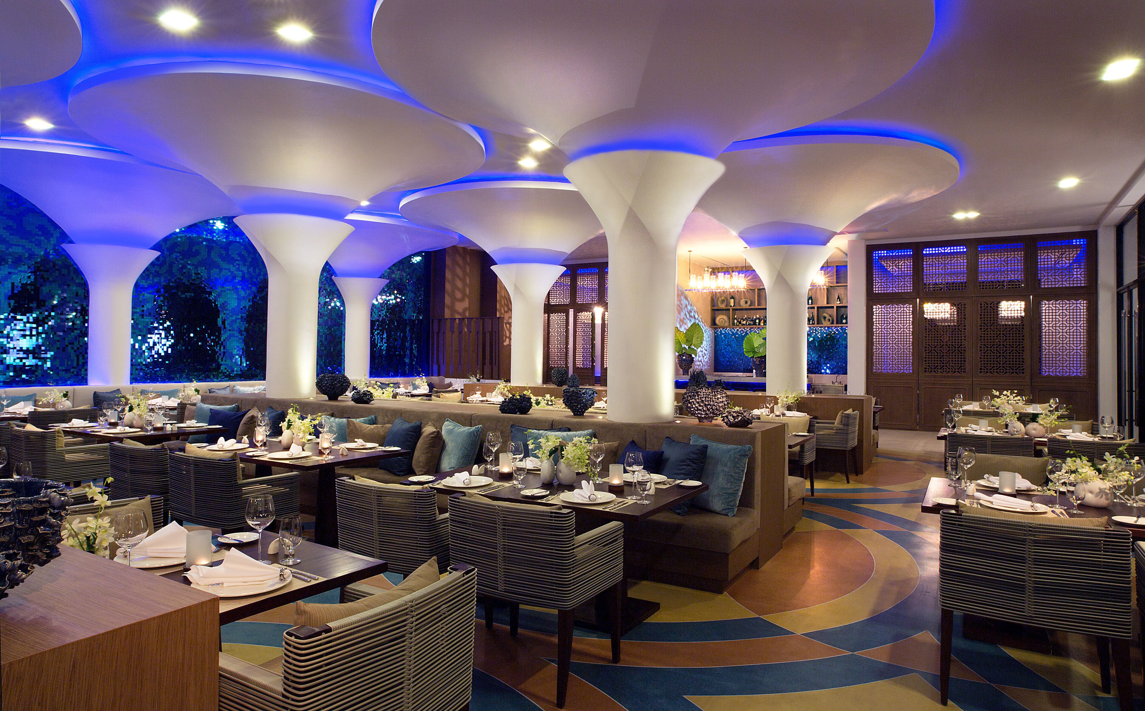 Ein Restaurant mit verschiedenen Essbereichen, darunter gemütliche Nischen und Tische, einem modernen Deckenbau und großen Fenstern.