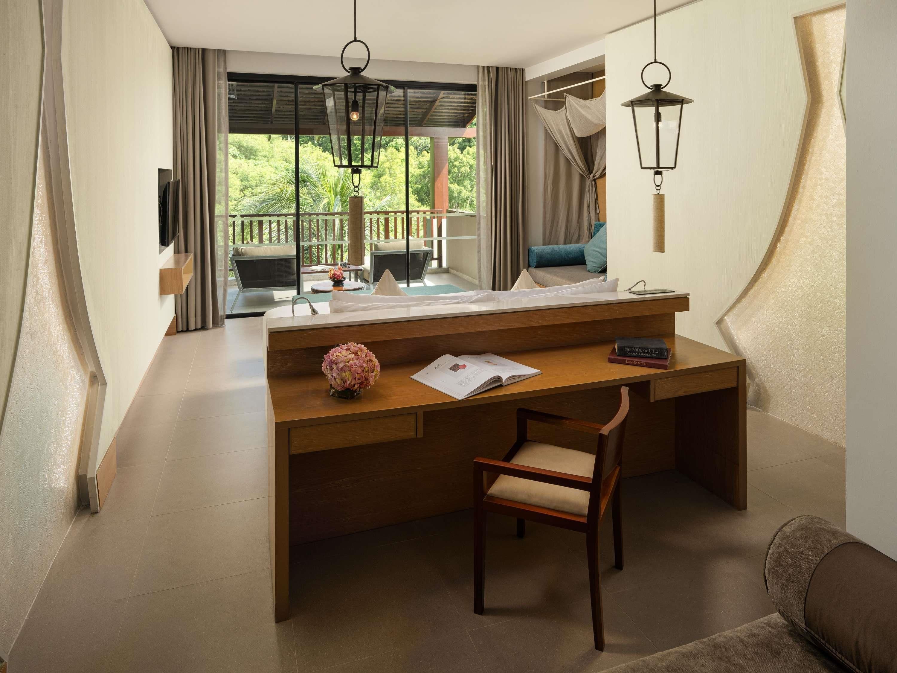 Une chambre d’hôtel avec un bureau en bois, une chaise, une grande fenêtre avec des rideaux, un balcon et une lampe suspendue.