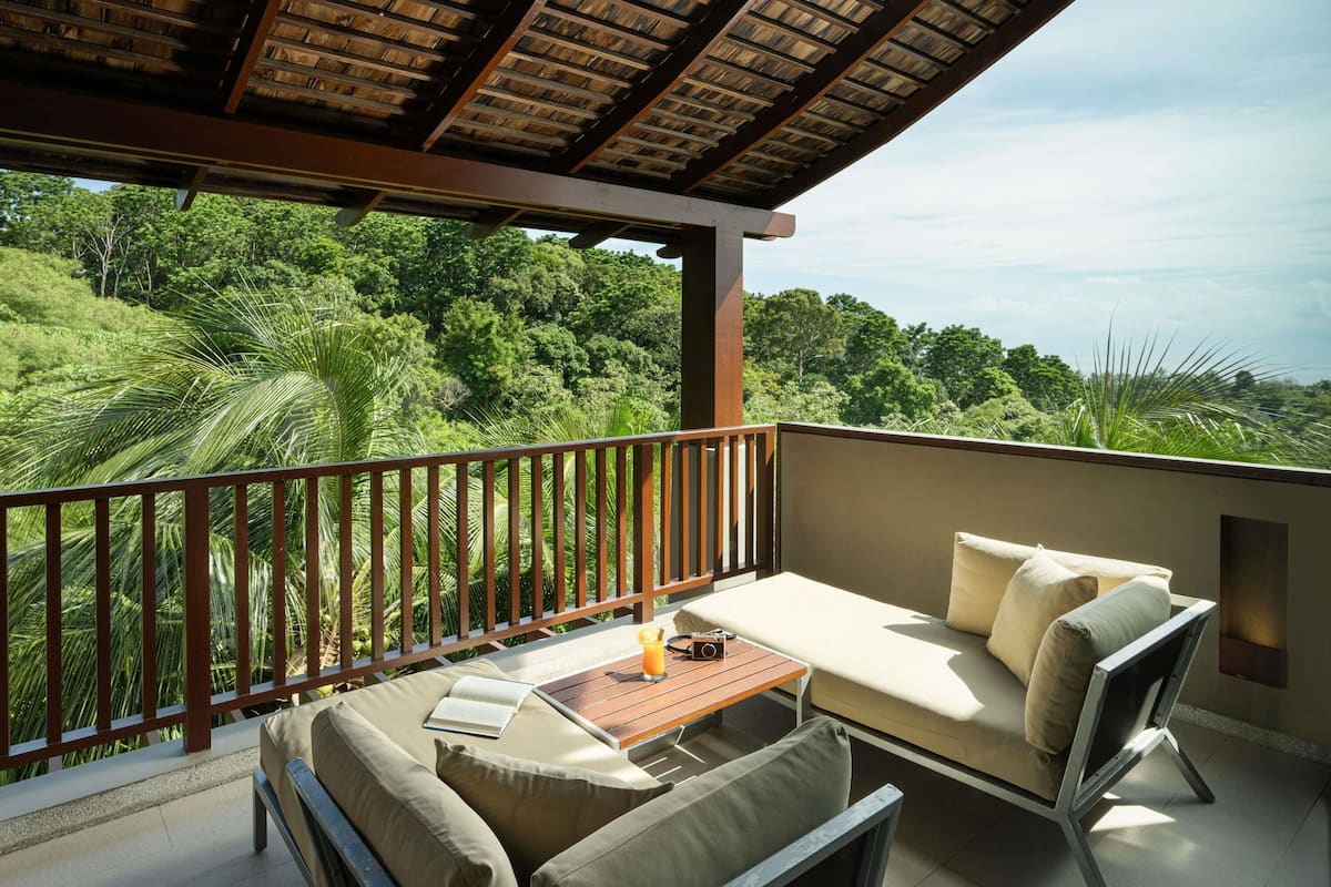 A balcony with a wooden ceiling, cushioned seating, a small table, and a view of lush greenery.
